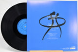 Vinyl record partially pulled from its blue sleeve, showing the black record, blue center label, and back cover artwork with white track listing text and a black abstract figure graphic on a bright blue background.
