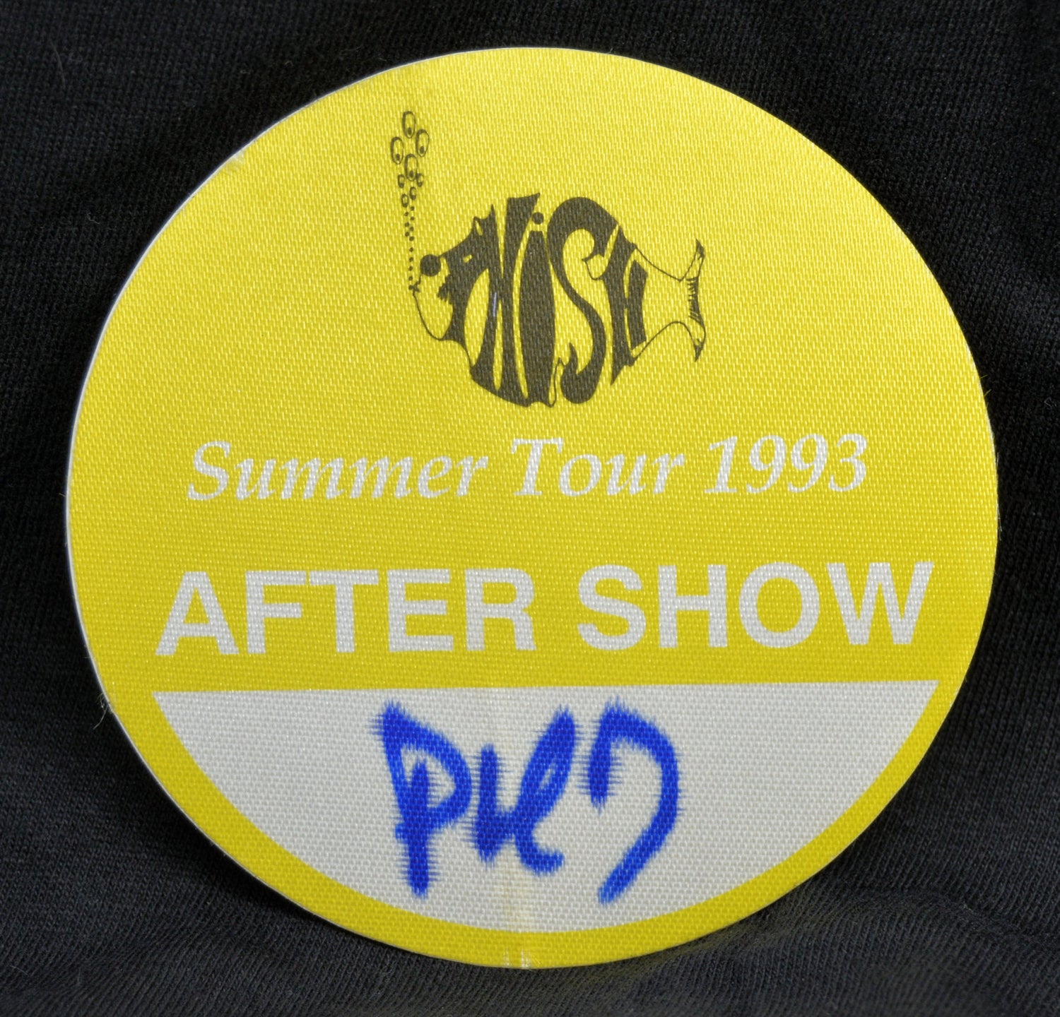 A round yellow backstage pass with a black fish logo and the text 'Nash Summer Tour 1993 AFTER SHOW', with a white curved section at the bottom containing blue handwritten text, shown on a dark fabric background.