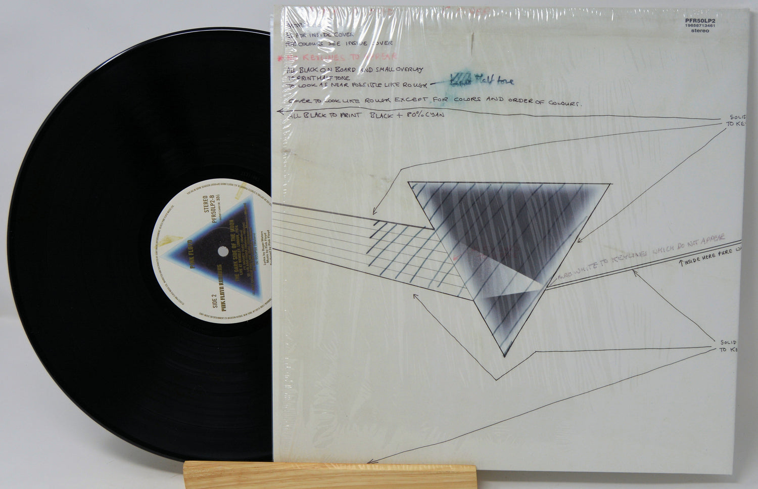A vinyl record partially pulled from its sealed white cover, displayed on a small wooden stand against a light gray background. The record is black with a blue-and-white center label, and the cover features a geometric triangle design.