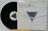 A vinyl record partially pulled from its sealed white cover, displayed on a small wooden stand against a light gray background. The record is black with a blue-and-white center label, and the cover features a geometric triangle design.