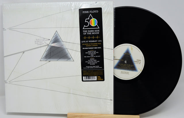 Pink Floyd 'The Dark Side of the Moon' 12-inch vinyl record shown in its shrink-wrapped outer sleeve with the black LP record partially pulled out, displayed on a small wooden stand against a light gray background.