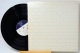 A vinyl record partially slid out of its cream-colored square album sleeve, which features a light blue brick pattern. The black record has a purple and white center label.