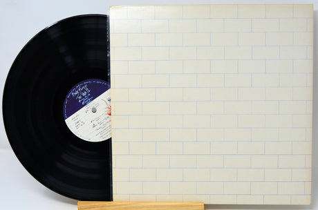 A vinyl record partially slid out of its cream-colored square album sleeve, which features a light blue brick pattern. The black record has a purple and white center label.