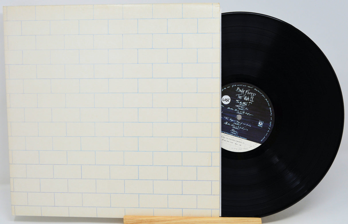 A vinyl record partially pulled from its sleeve, displayed on a small wooden stand against a light gray background. The sleeve features a white brick pattern with thin blue lines, and the black vinyl disc has a white and black center label.