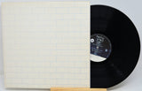 A vinyl record partially pulled from its sleeve, displayed on a small wooden stand against a light gray background. The sleeve features a white brick pattern with thin blue lines, and the black vinyl disc has a white and black center label.
