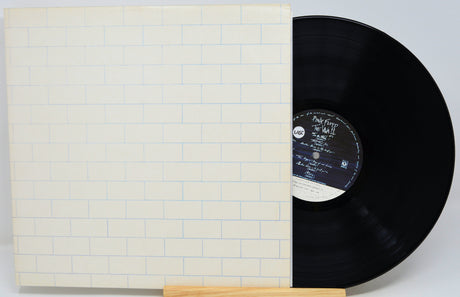 A vinyl record partially pulled from its sleeve, displayed on a small wooden stand against a light gray background. The sleeve features a white brick pattern with thin blue lines, and the black vinyl disc has a white and black center label.