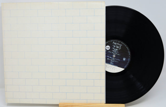 A vinyl record partially pulled from its sleeve, displayed on a small wooden stand against a light gray background. The sleeve features a white brick pattern with thin blue lines, and the black vinyl disc has a white and black center label.