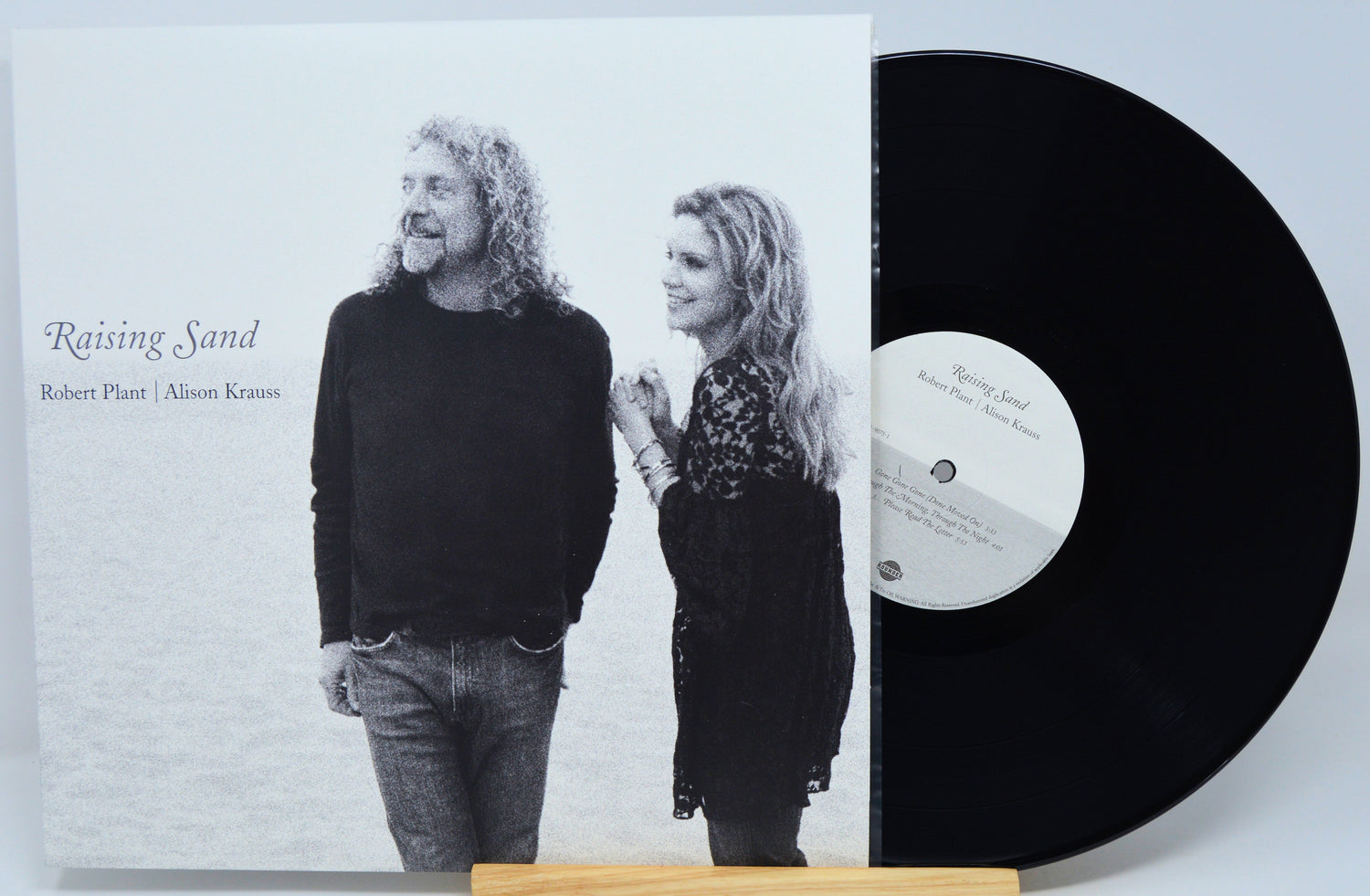 A vinyl record partially pulled from its sleeve, showing the album cover for 'Raising Sand' by Robert Plant and Alison Krauss. The sleeve features a black-and-white photo of the couple, and the black record is visible on a small wooden stand against a light gray background.