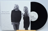 A vinyl record partially pulled from its sleeve, showing the album cover for 'Raising Sand' by Robert Plant and Alison Krauss. The sleeve features a black-and-white photo of the couple, and the black record is visible on a small wooden stand against a light gray background.
