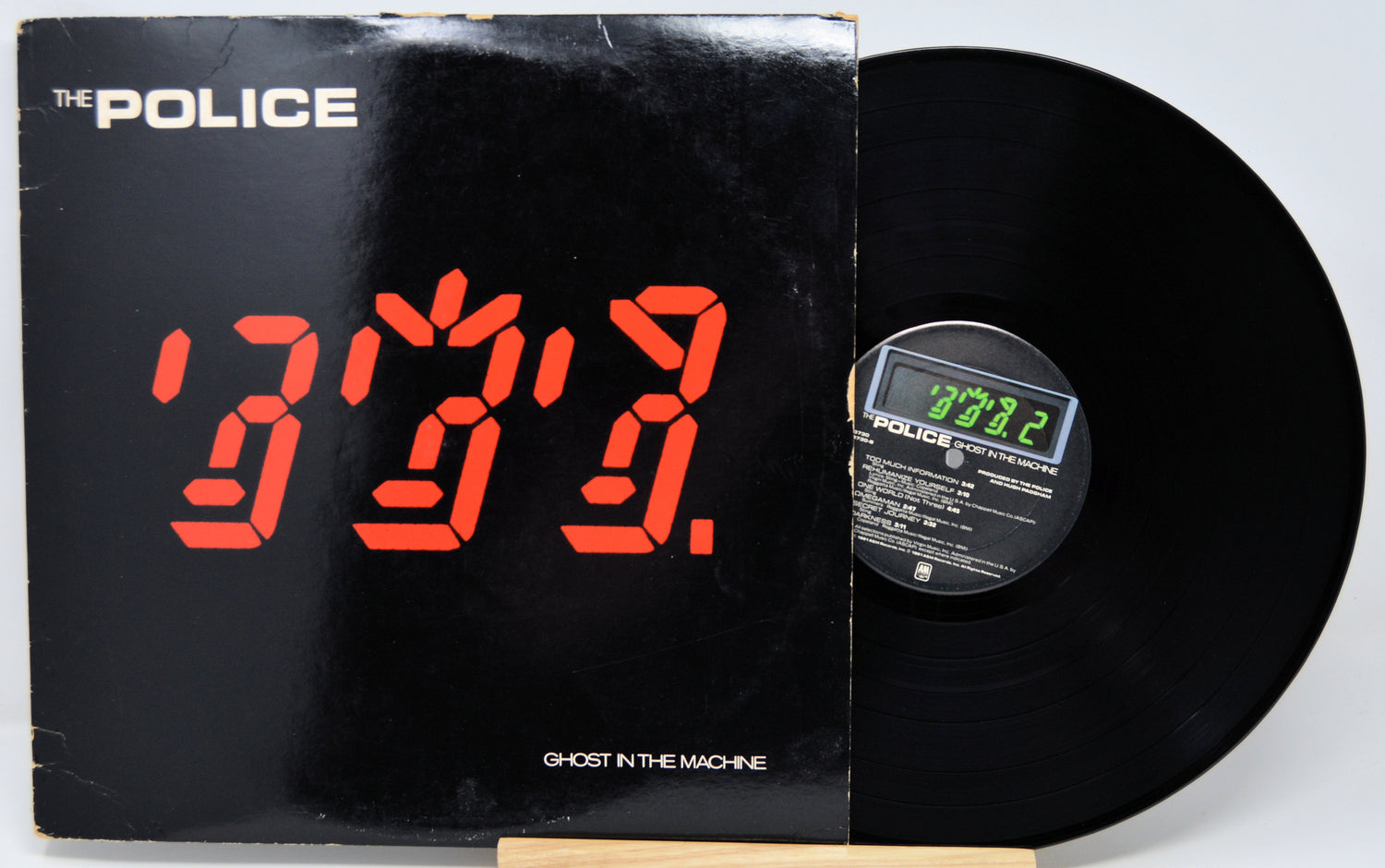 The Police 'Ghost in the Machine' vinyl record LP with its black cover featuring a red digital clock display, shown next to the black record on a light gray background.