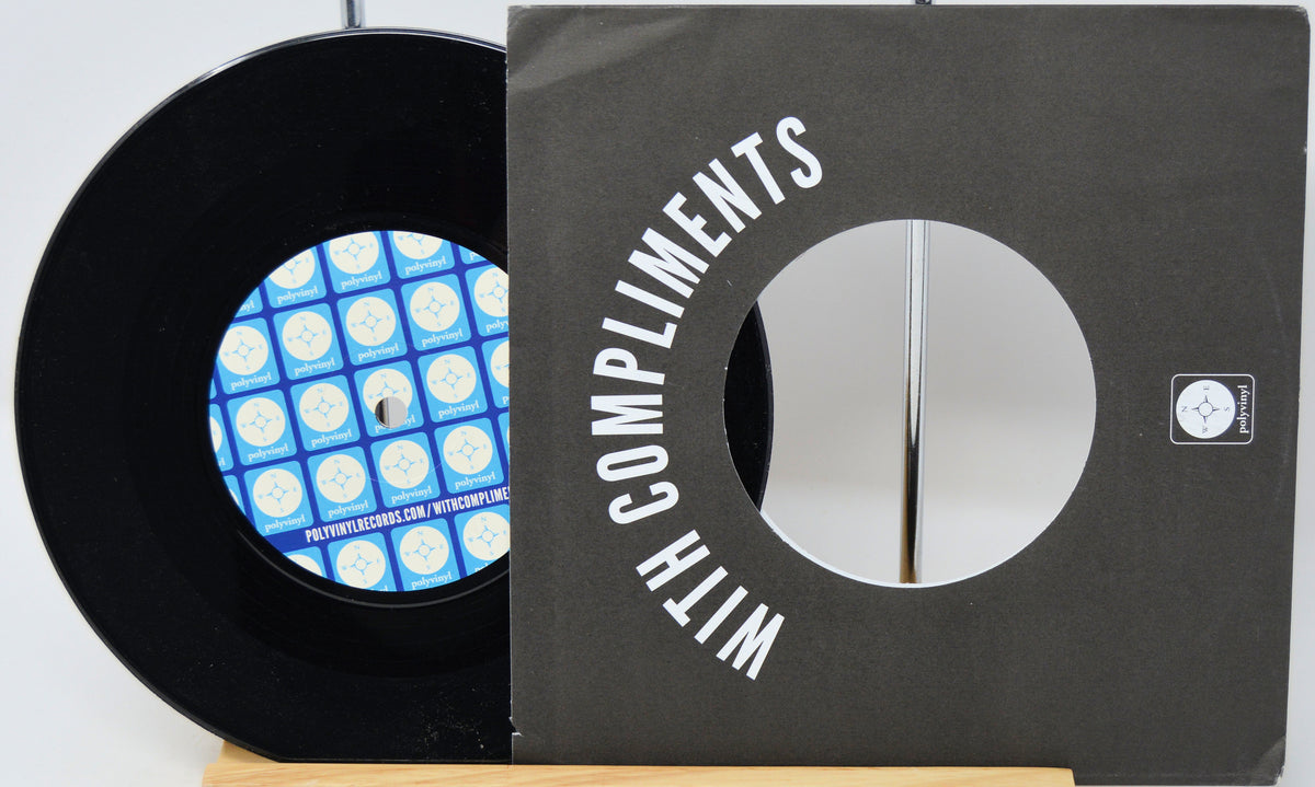 A black vinyl record partially pulled from a dark gray sleeve with large white text reading 'WITH COMPLIMENTS.' The record label has a blue and white geometric pattern, and a small square logo is visible on the sleeve.