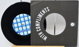 A black vinyl record partially pulled from a dark gray sleeve with large white text reading 'WITH COMPLIMENTS.' The record label has a blue and white geometric pattern, and a small square logo is visible on the sleeve.