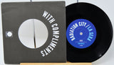 A black vinyl record partially pulled from its sleeve, showing the black cover with white text reading 'WITH COMPLIMENTS' and the blue record label reading 'RADIATION CITY / LA SERA' and 'JUCY'.