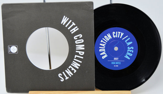 A black vinyl record partially pulled from its sleeve, showing the black cover with white text reading 'WITH COMPLIMENTS' and the blue record label reading 'RADIATION CITY / LA SERA' and 'JUCY'.