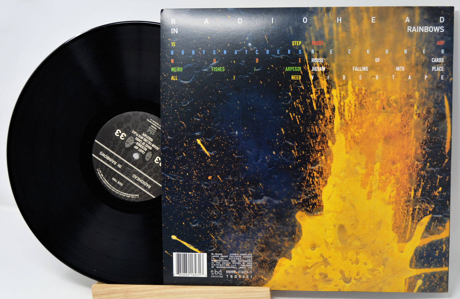 Radiohead 'Rainbows' vinyl record shown with the black LP partially pulled from the back cover, which features a dark blue and bright yellow-orange abstract splash design.