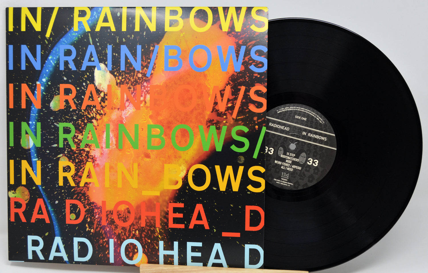 A vinyl record partially pulled from its sleeve, showing a black disc and a square album cover with a colorful abstract design and repeated rainbow text reading 'IN RAINBOWS'.
