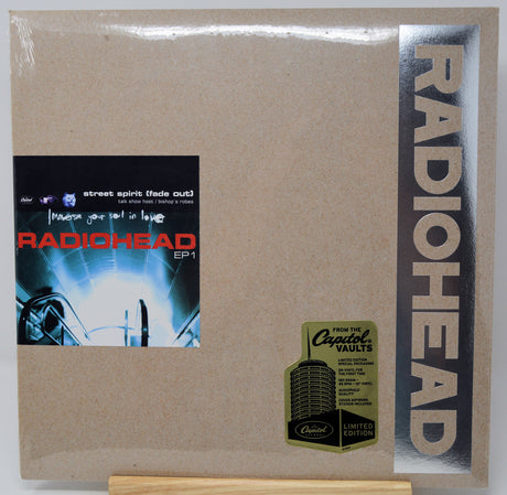 Radiohead 'EP1' vinyl record cover in its original shrink wrap, displayed on a small wooden stand. The cover features a textured brown kraft-paper background, a vertical black band on the right with 'RADIOHEAD' in large silver letters, and a black square album art label on the left.