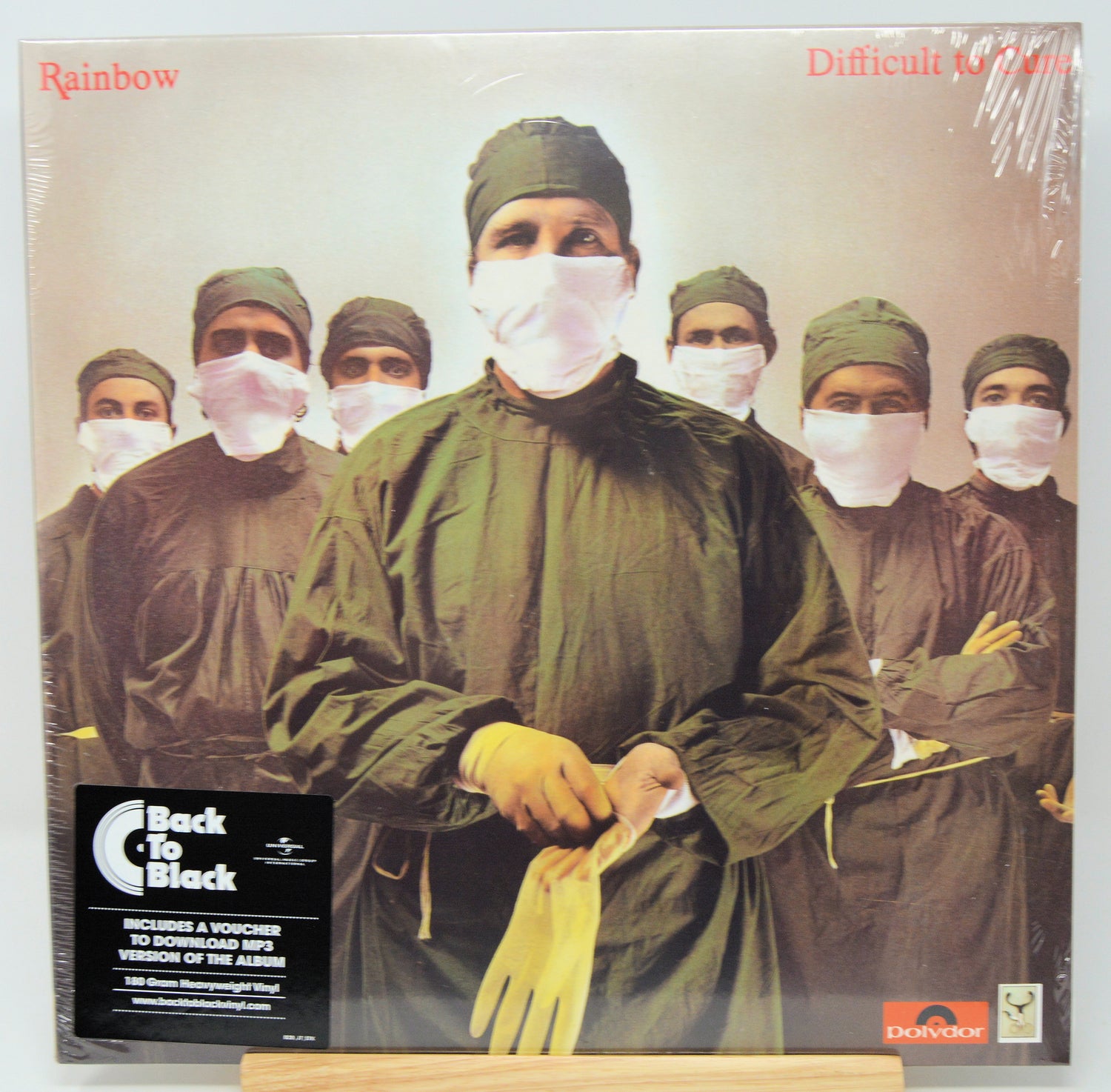The Back To Black album 'Rainbow' vinyl LP cover, sealed in its original plastic shrink wrap and displayed on a wooden stand. The cover art features a group of surgeons in green uniforms and white face masks, with the title 'Rainbow' in red in the top left corner and a black 'Back To Black' sticker on the bottom left.