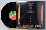 A vinyl record partially pulled from its sleeve, showing the back of the album case with fiery orange flames and track listing text on a black background. The black record itself is visible on the left, resting on a light wood surface against a white backdrop.