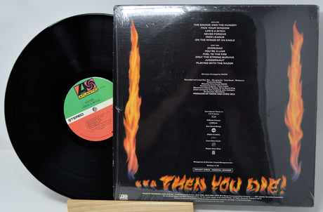 A vinyl record partially pulled from its sleeve, showing the back of the album case with fiery orange flames and track listing text on a black background. The black record itself is visible on the left, resting on a light wood surface against a white backdrop.