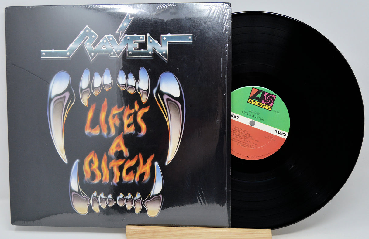 A vinyl record album for the band Raven's 'Life's A Bitch' on two records, with the black LP disc partially pulled from the sleeve. The sleeve features a black background with a graphic of metallic silver fangs and orange text.