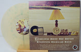 A vinyl record album shown with its printed cover art and a translucent pale yellow record partially visible behind it. The cover features a scenic landscape and a table scene with a lamp, clock, books, and a phone, with track listing text at the bottom.