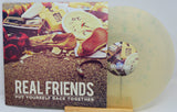Real Friends vinyl record album with the LP partially pulled from the sleeve, showing a translucent pale green record and cover art featuring a collage of objects on a textured ground with the title 'REAL FRIENDS PUT YOURSELF BACK TOGETHER'.