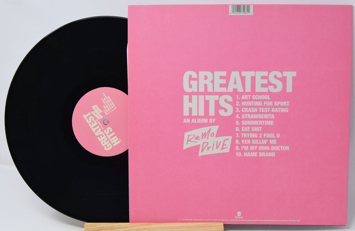 A vinyl record partially pulled from its pink sleeve, showing the black record and a pink center label with the text 'The Sun Is Still Rising.' The sleeve has a large pink cover with white text reading 'GREATEST HITS' and a tracklist, photographed on a light background.