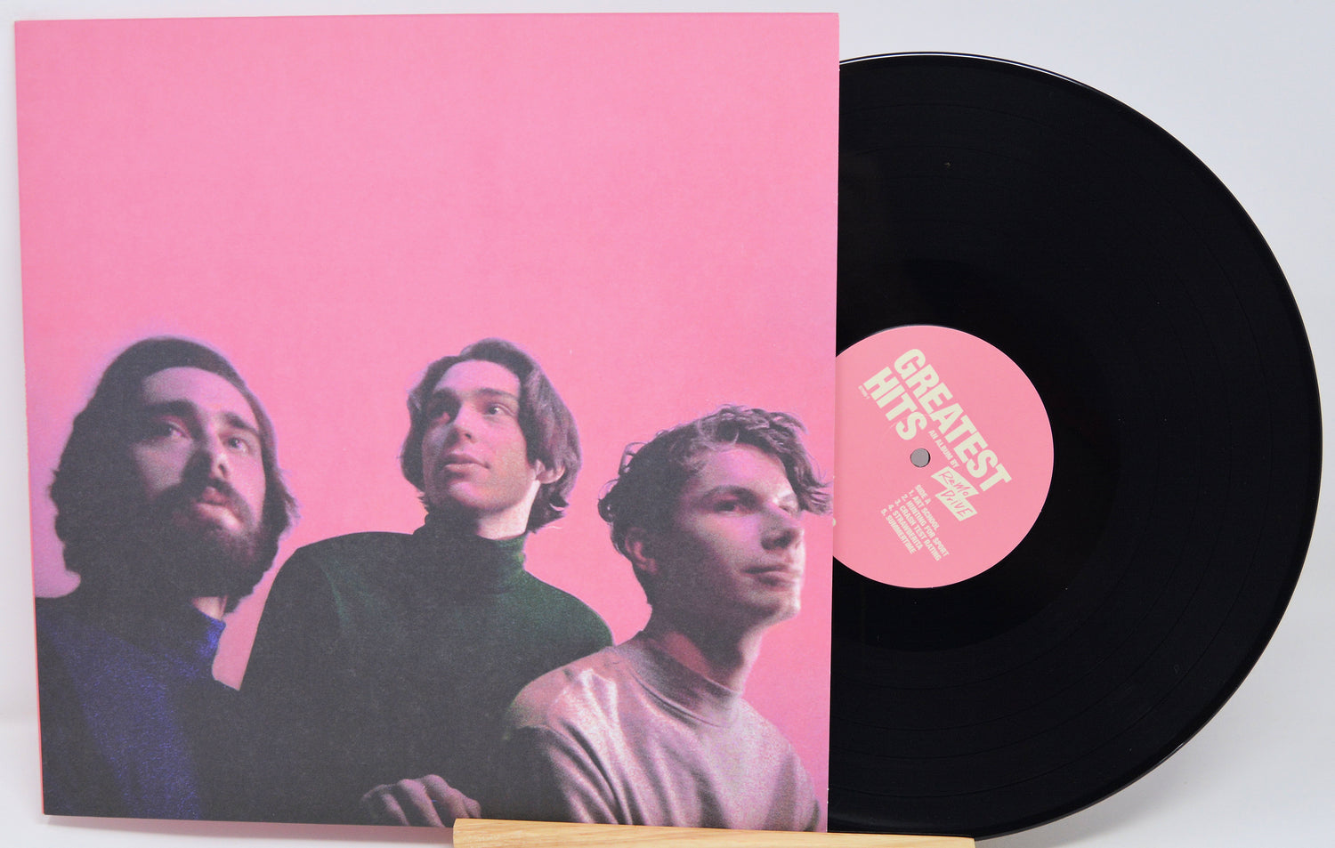A vinyl record partially slid out of its sleeve, showing a black record with a pink center label and a pink album cover featuring three men against a pink background.