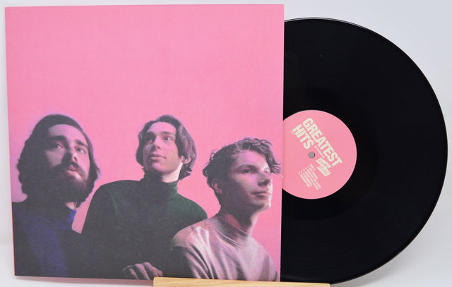 A vinyl record partially slid out of its sleeve, showing a black record with a pink center label and a pink album cover featuring three men against a pink background.