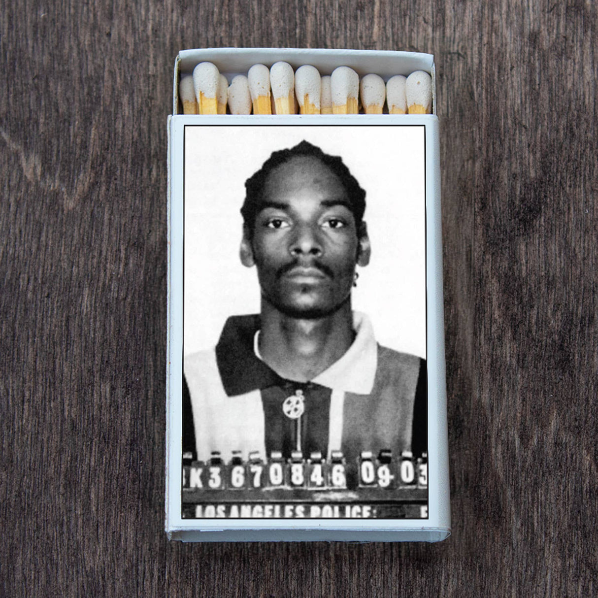 A retro-style matchbox featuring a black and white mugshot on the cover, shown on a wooden surface.