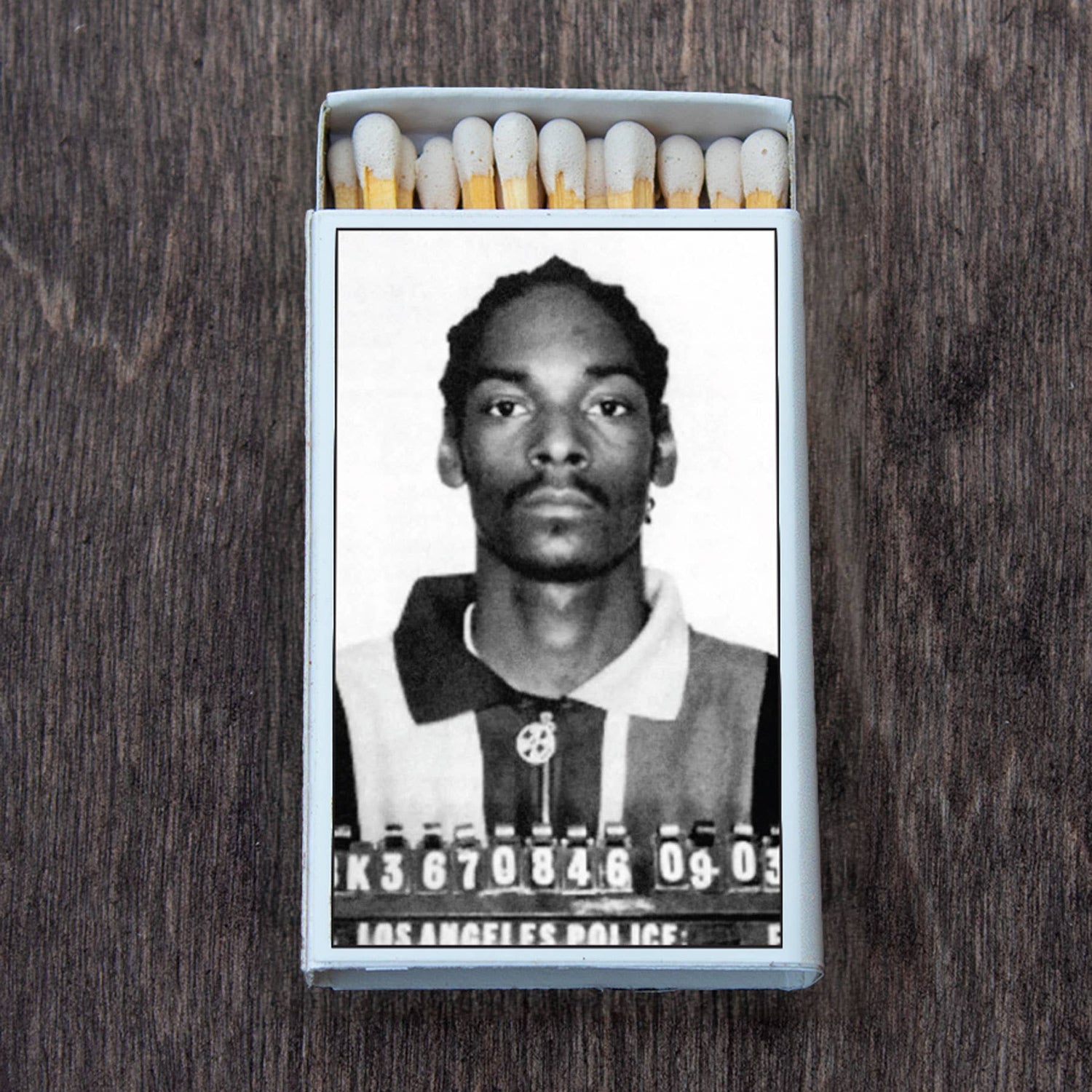 A retro-style matchbox featuring a black and white mugshot on the cover, shown on a wooden surface.