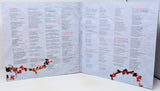 An open three-panel accordion-style booklet or program spread on a white surface, printed with many small text blocks and decorated with red and orange floral garlands along the bottom corners.