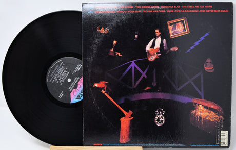 A vinyl record partially pulled from its illustrated black album cover, photographed on a plain white background. The cover artwork is dark with purple, orange, and red graphic elements, and the black record sleeve is visible behind the black vinyl disc.