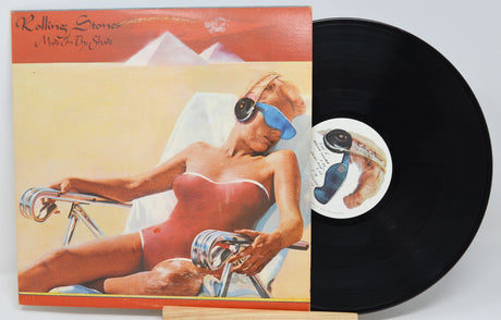 The image shows a used vinyl record of 'Rolling Stones - Made In The Shade' with a picture of a female figure in a bikini, sunglasses, and a hat, lounging in a chair.