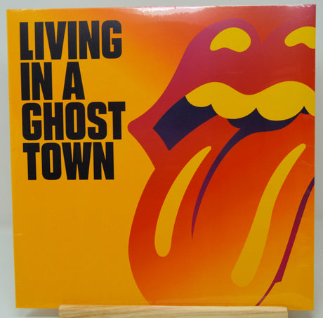 Rolling Stones 'Living in a Ghost Town' vinyl record album cover, featuring the band's iconic tongue logo in yellow, orange, and purple on a bright yellow-orange background, with the album title in bold black text. The vinyl is displayed on a light-colored wooden stand against a neutral light gray background.