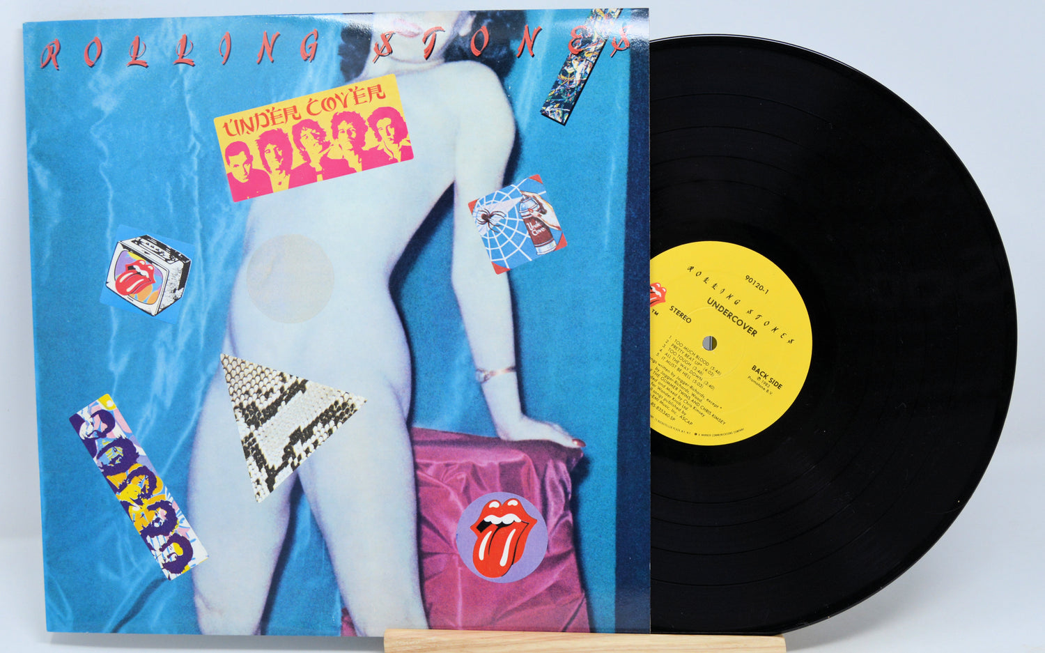 This image features a vintage vinyl record of "Rolling Stones - Under Cover," which is housed in a standard-sized album cover. The cover displays the band's iconic logo and a provocative visual of a member in a compromising position, encapsulating the band's edgy image.