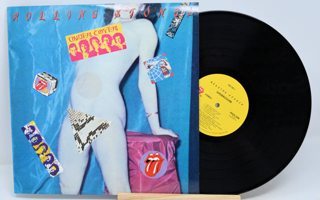 This image features a vintage vinyl record of "Rolling Stones - Under Cover," which is housed in a standard-sized album cover. The cover displays the band's iconic logo and a provocative visual of a member in a compromising position, encapsulating the band's edgy image.