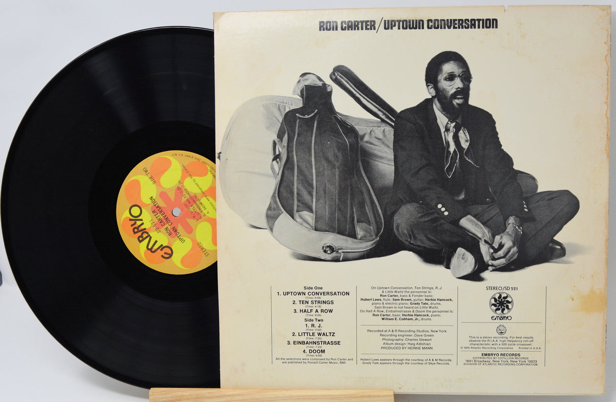 Ron Carter / Uptown Conversation vinyl record album shown with the black LP partially pulled from the cover, photographed on a white background. The cover features a black-and-white portrait of Ron Carter sitting cross-legged with a guitar, and the record label shows OVO branding.
