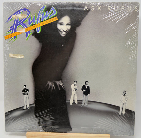 A vinyl record album for 'Ask Rufus' by Rufus, sealed in clear plastic shrink wrap and displayed on a wooden stand. The cover features a black and white photo of a smiling woman in a long dress, with four small figures standing on the floor, and stylized blue and orange 'Rufus' logo text.