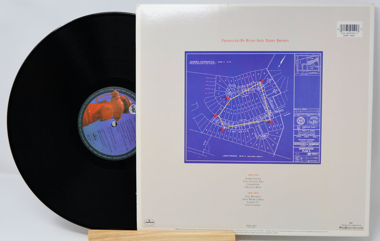 A vinyl record partially pulled from its sleeve, showing the black record with a blue and orange label next to the cream-colored cover featuring a blue architectural blueprint design.