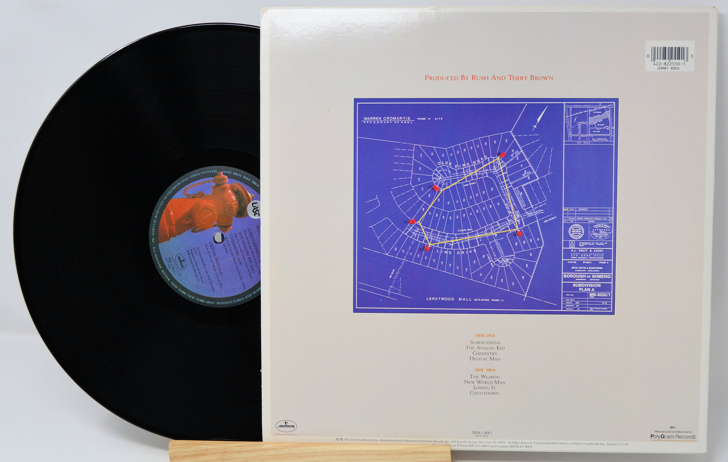 A vinyl record partially pulled from its sleeve, showing the black record with a blue and orange label next to the cream-colored cover featuring a blue architectural blueprint design.