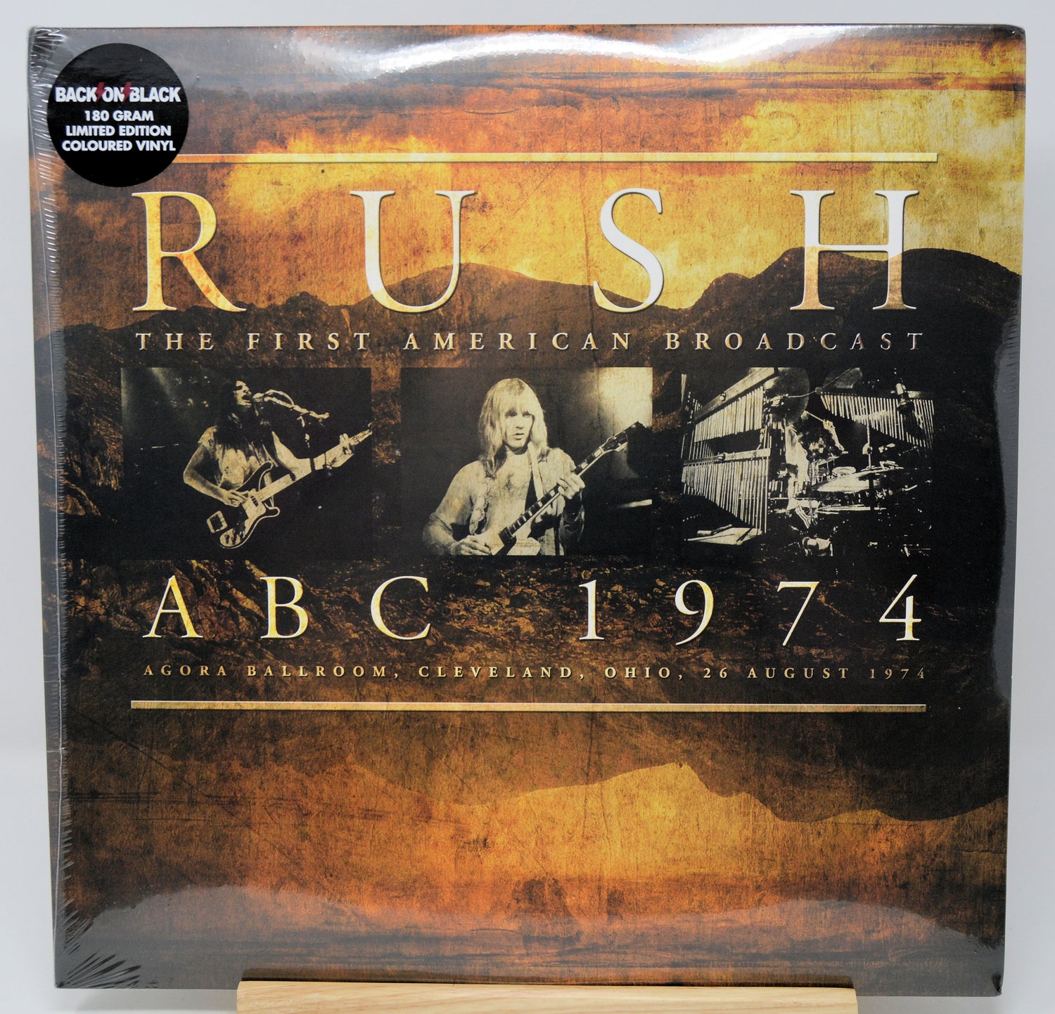 Rush - The First American Broadcast ABC 1974 vinyl record LP cover, shown in shrink wrap on a wooden display stand. The cover features a textured brown and gold design with black-and-white photos of the band members and a black 'Back on Black' 12-inch colored vinyl sticker in the top left corner.