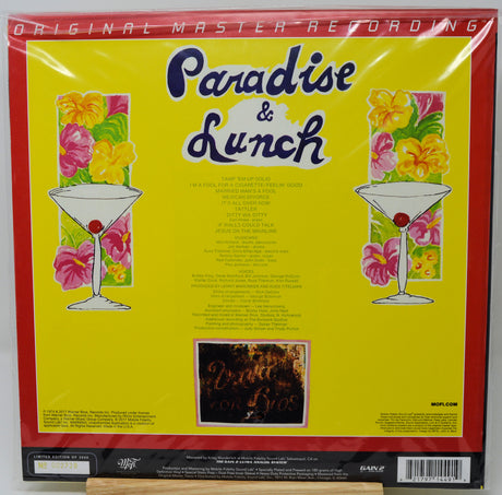The back cover of the vinyl record 'Paradise & Lunch' by Paradise Lost, sealed in its original plastic shrink wrap. The cover features a bright yellow background with a thick red border, decorated with two colorful floral panels flanking martini glasses, and the band's name in stylized blue lettering at the top.