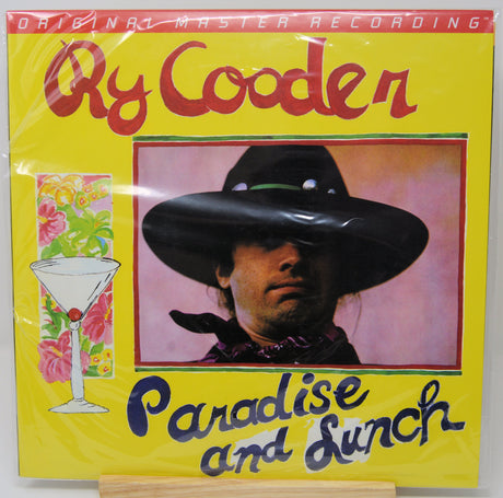A Ry Cooder 'Paradise and Sunch' vinyl record album cover, sealed in its original plastic shrink wrap and displayed on a small wooden stand. The cover features a bright yellow background with red and blue text, and a central portrait of the artist in a black hat.