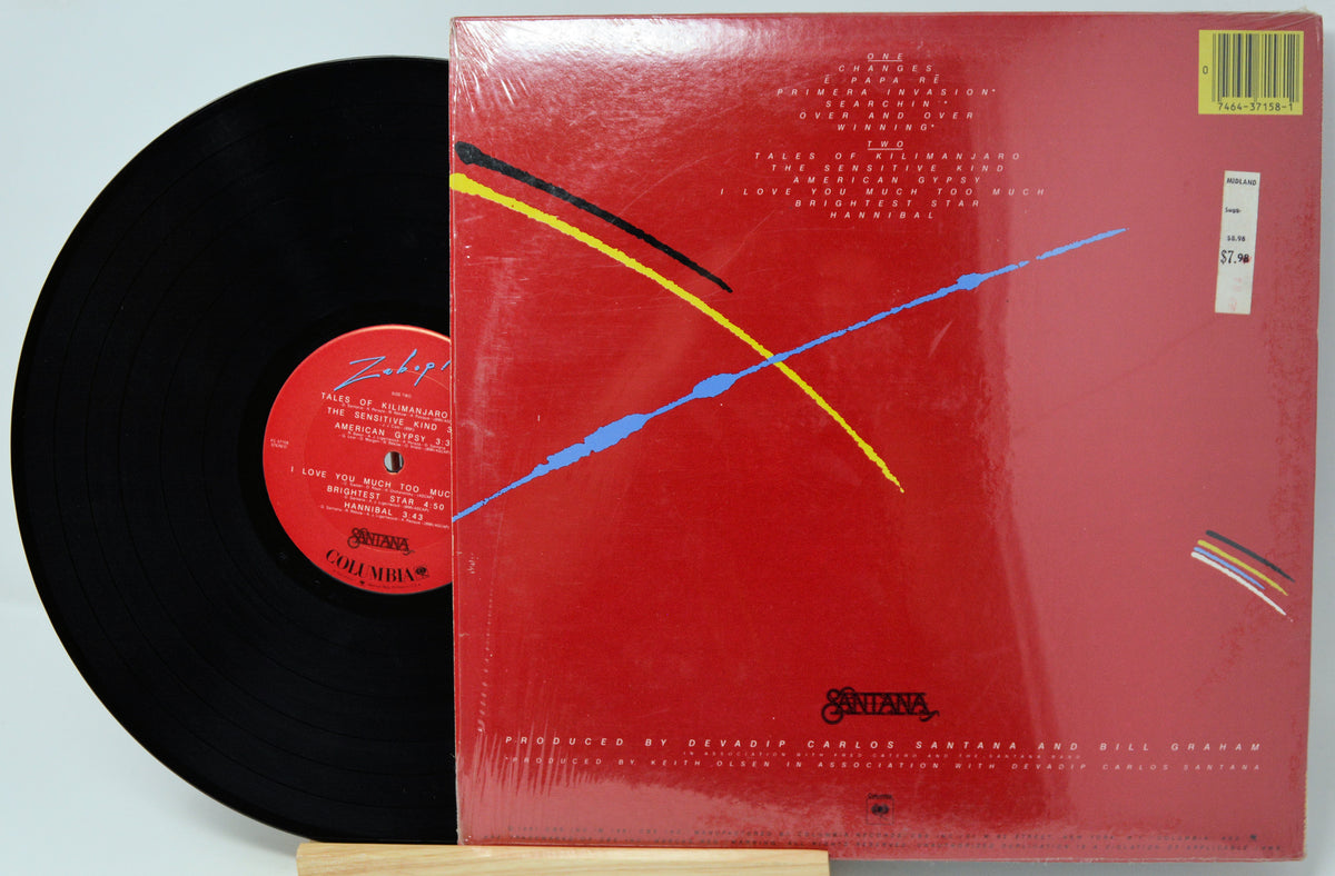 A vinyl record partially pulled from its red sleeve, photographed on a light gray background. The record is black with a red center label, and the sleeve features abstract blue, yellow, and black line art on a red background.