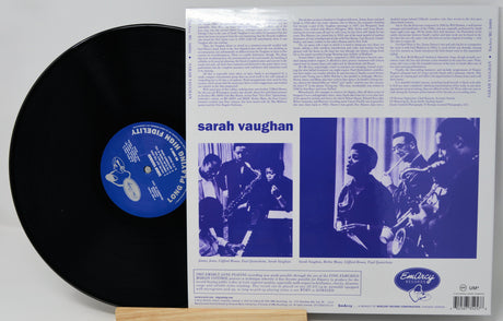 A vinyl record partially pulled from its back cover sleeve, showing the black record with a blue center label next to the printed album back cover for Sarah Vaughan's 'Love Is A Drug'.