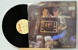 Vinyl record with cover featuring a couple holding a framed photo, on a light gray background.