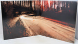 A square art print or photo print displayed standing open against a white background, showing a moody forest road scene with long exposure light trails and dark trees.