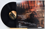 Vinyl record album shown with the black record partially pulled from the sleeve, photographed on a light background. The sleeve artwork features a dark forest scene with a house and tracklist text.